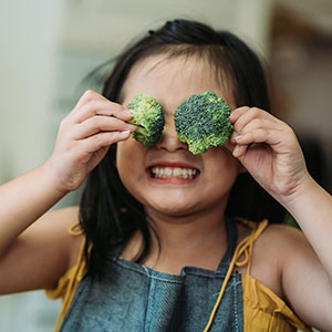 Child holds broccoli over her eyes